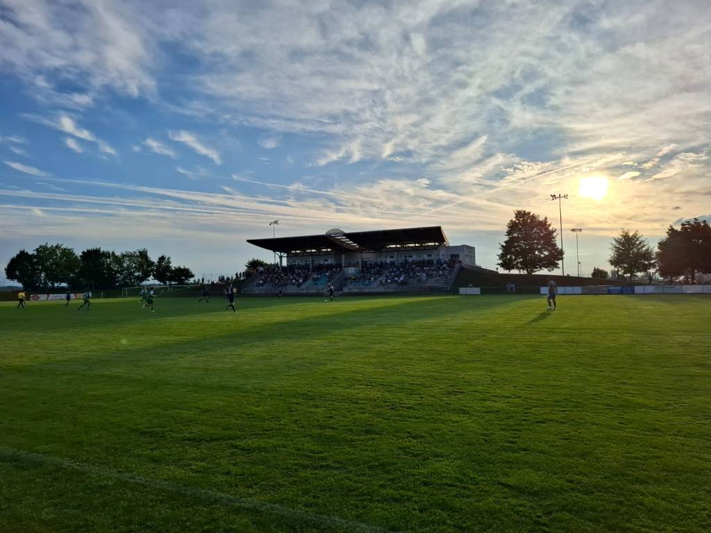 Fußballspiel auf einem Stadionrasen mit Zuschauern im Hintergrund unter einem klaren Himmel bei Sonnenuntergang.